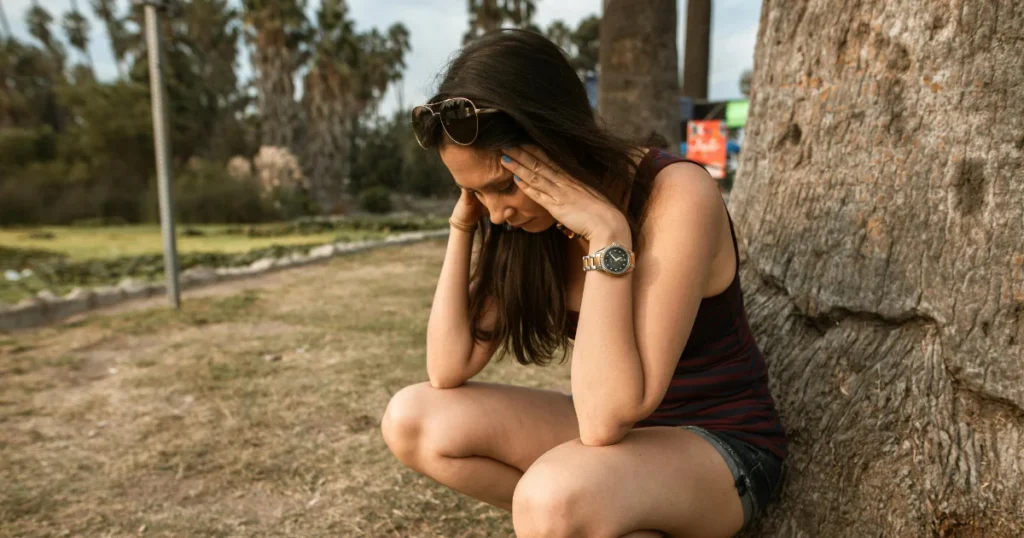 Uma mulher segurando a cabeça com as duas mãos e de olhos fechados sentada em uma arvore